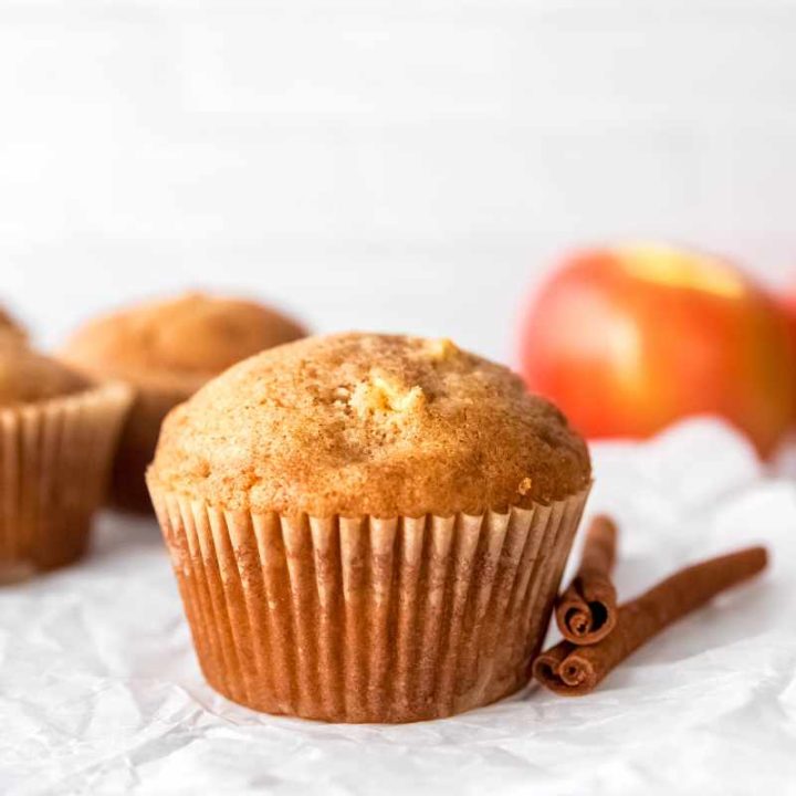 A perfectly domed apple cinnamon muffin rests on a parchment-lined cooling rack with two cinnamon sticks beside it.