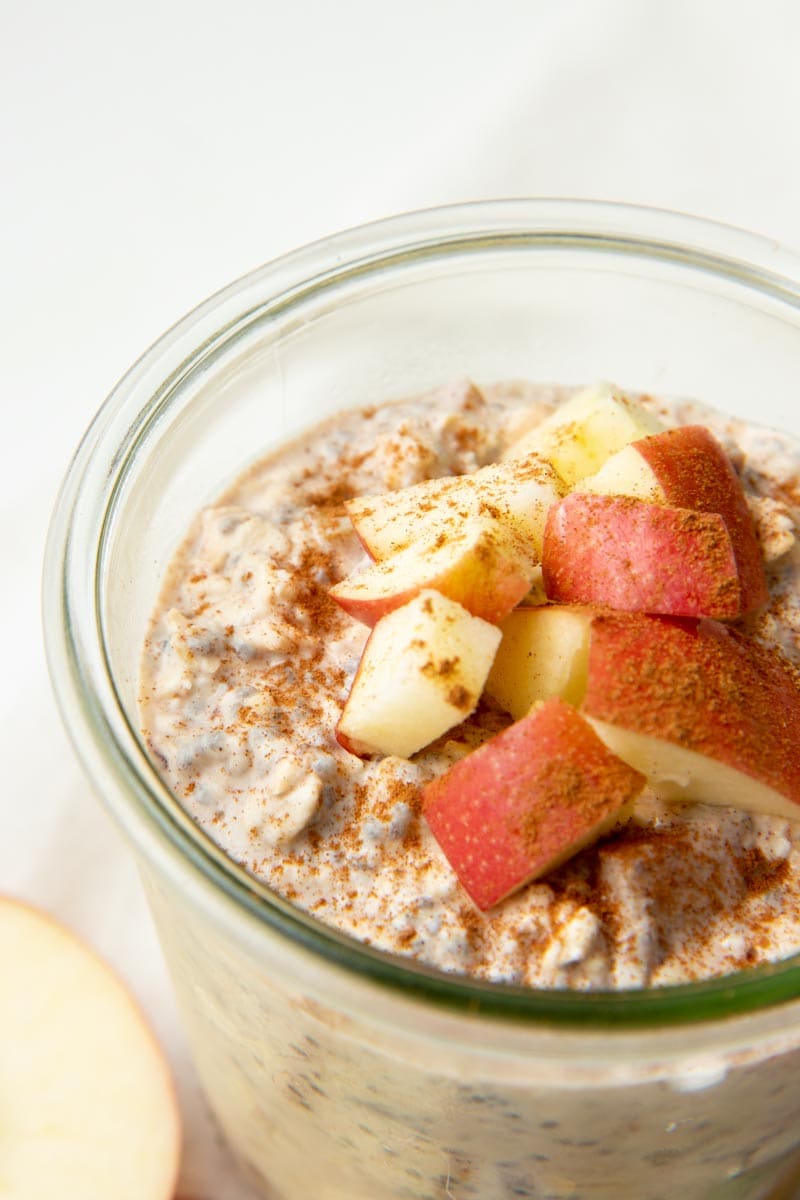 Tight view of apples and cinnamon topping a glass jar of overnight oats on a white background.