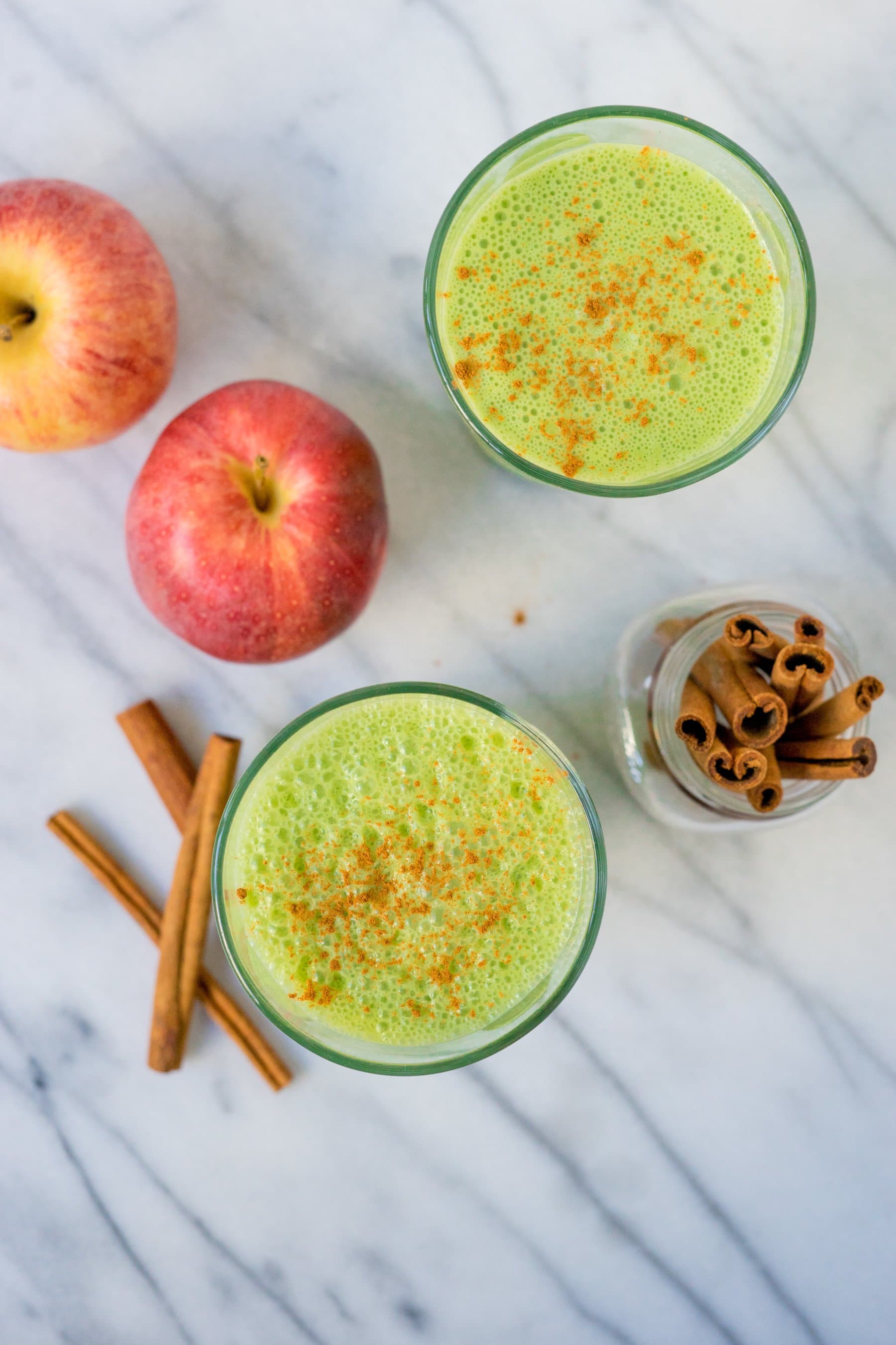 Overhead view of two glasses of green smoothie with apples and cinnamon sticks surrounding them