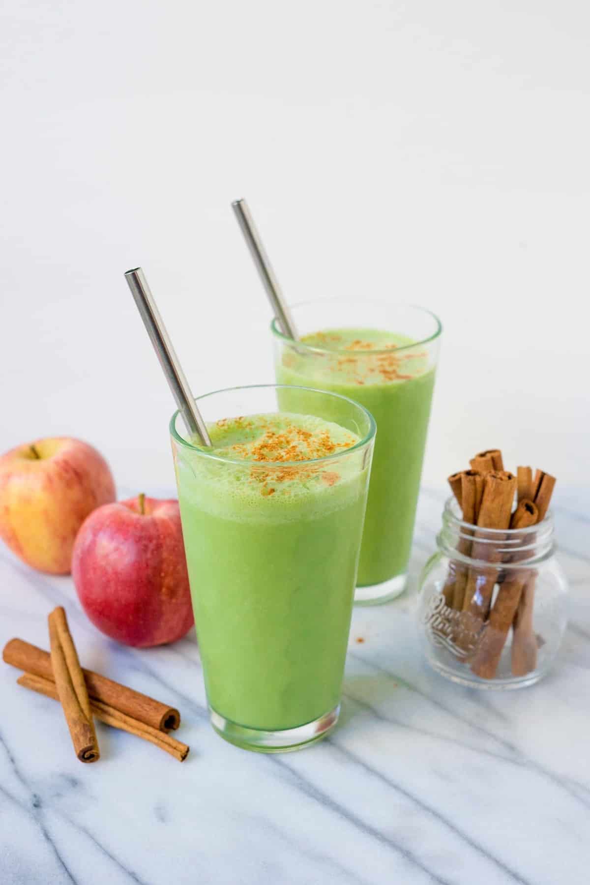 Two glasses of bright green smoothie with straws on white background with apples and cinnamon sticks garnish