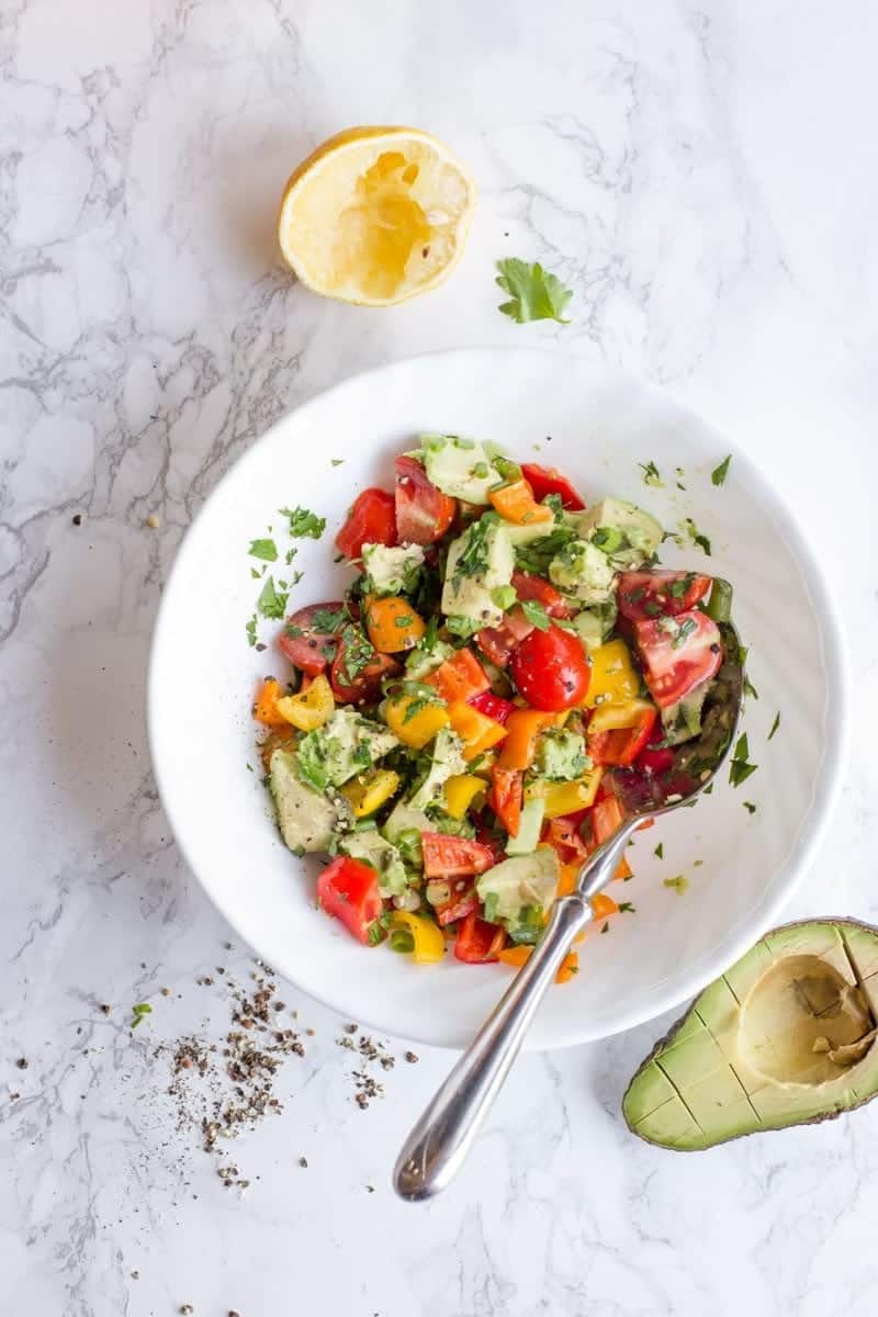 Avocado Bell Pepper Salad sits in a white bowl on a white background. A Lemon sits nearby.