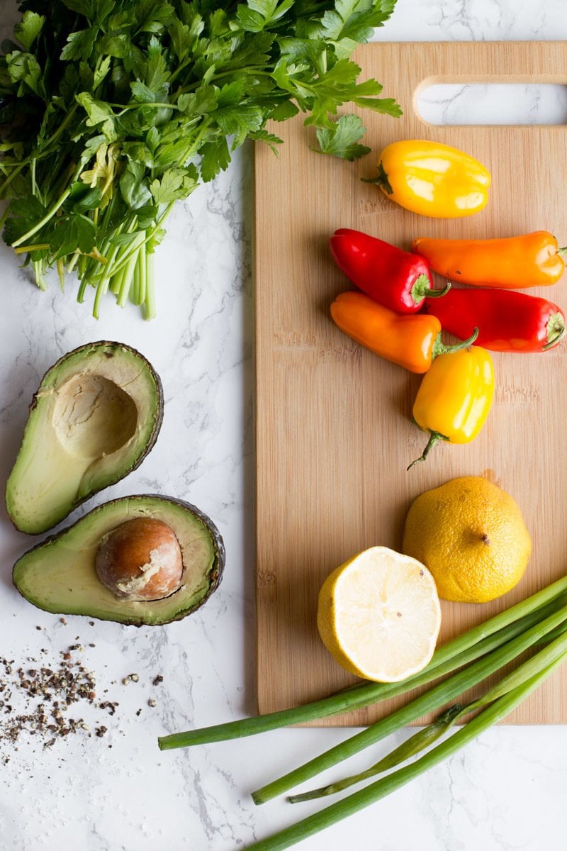 peppers, onion, lemon, cilantro and avocado sit on a wooden cutting board.