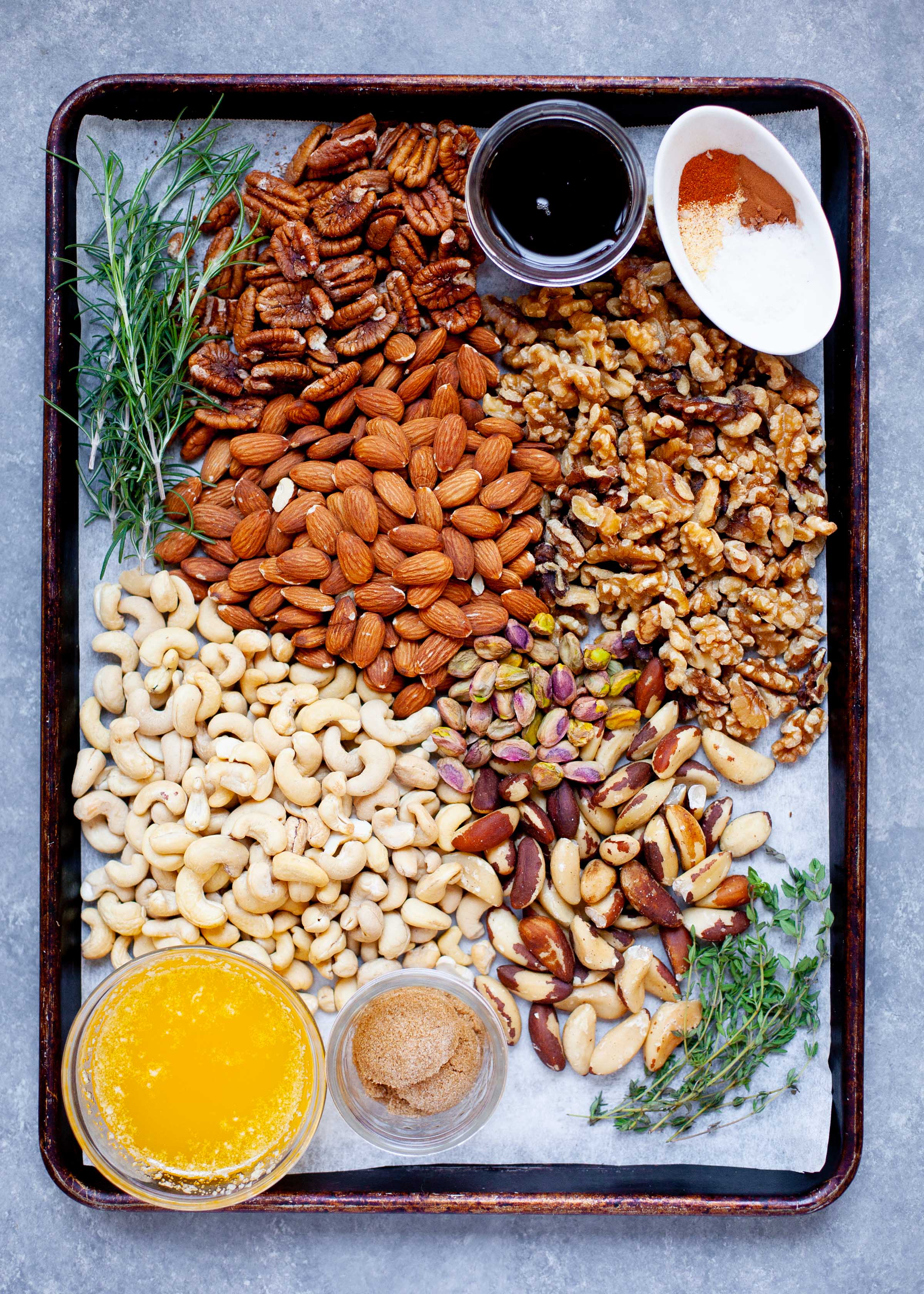 Ingredients for Maple Rosemary Bar Nuts on a white background - various nuts, rosemary, melted butter, brown sugar, rosemary, and spices