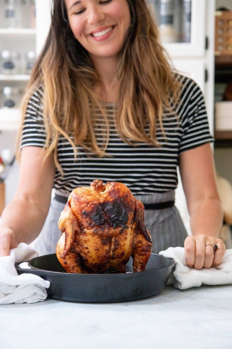 Grilled Beer Can Chicken standing up in a cast iron skillet, with a smiling woman in the background