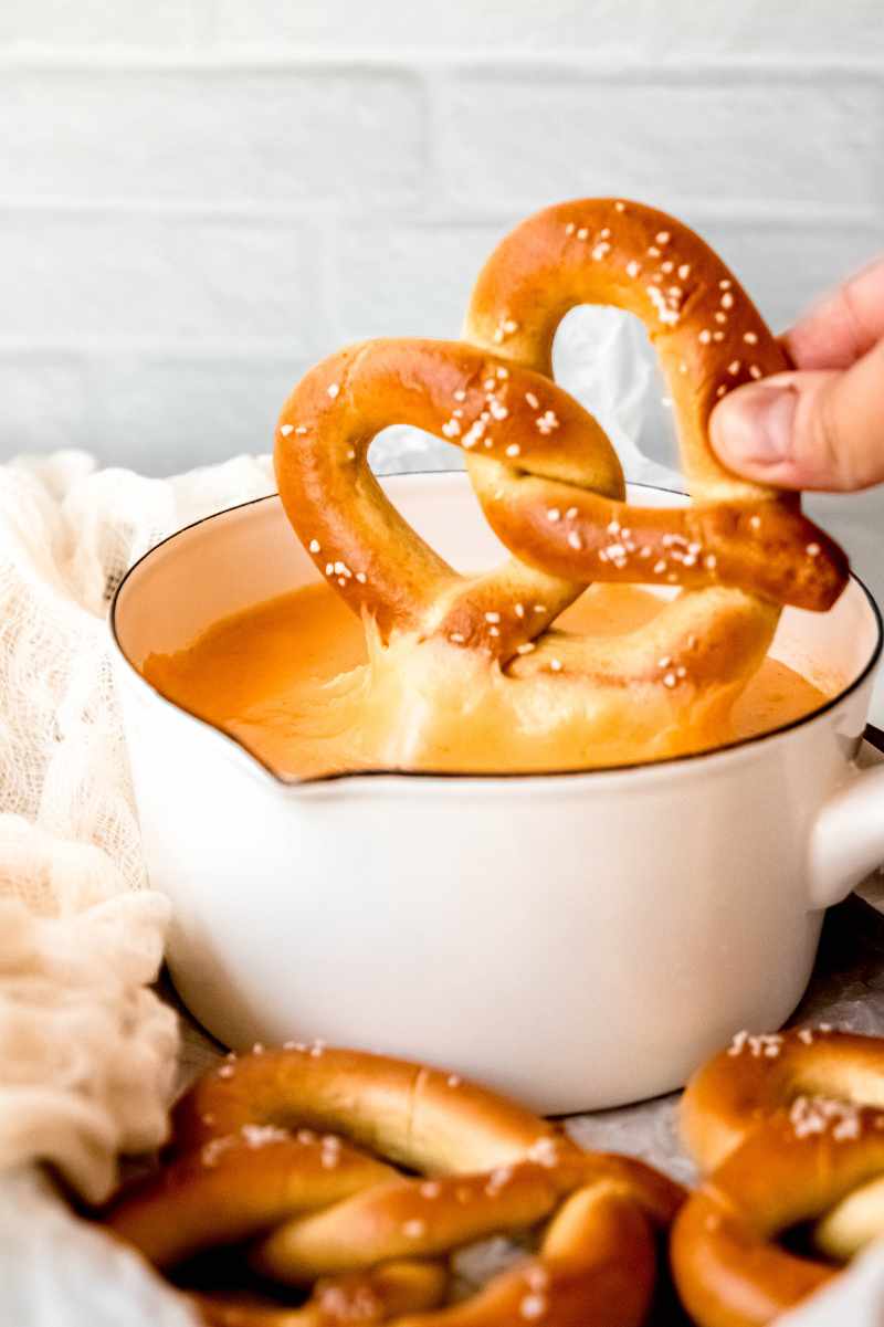 A hand dunks a pretzel into a small pot of beer cheese dip.