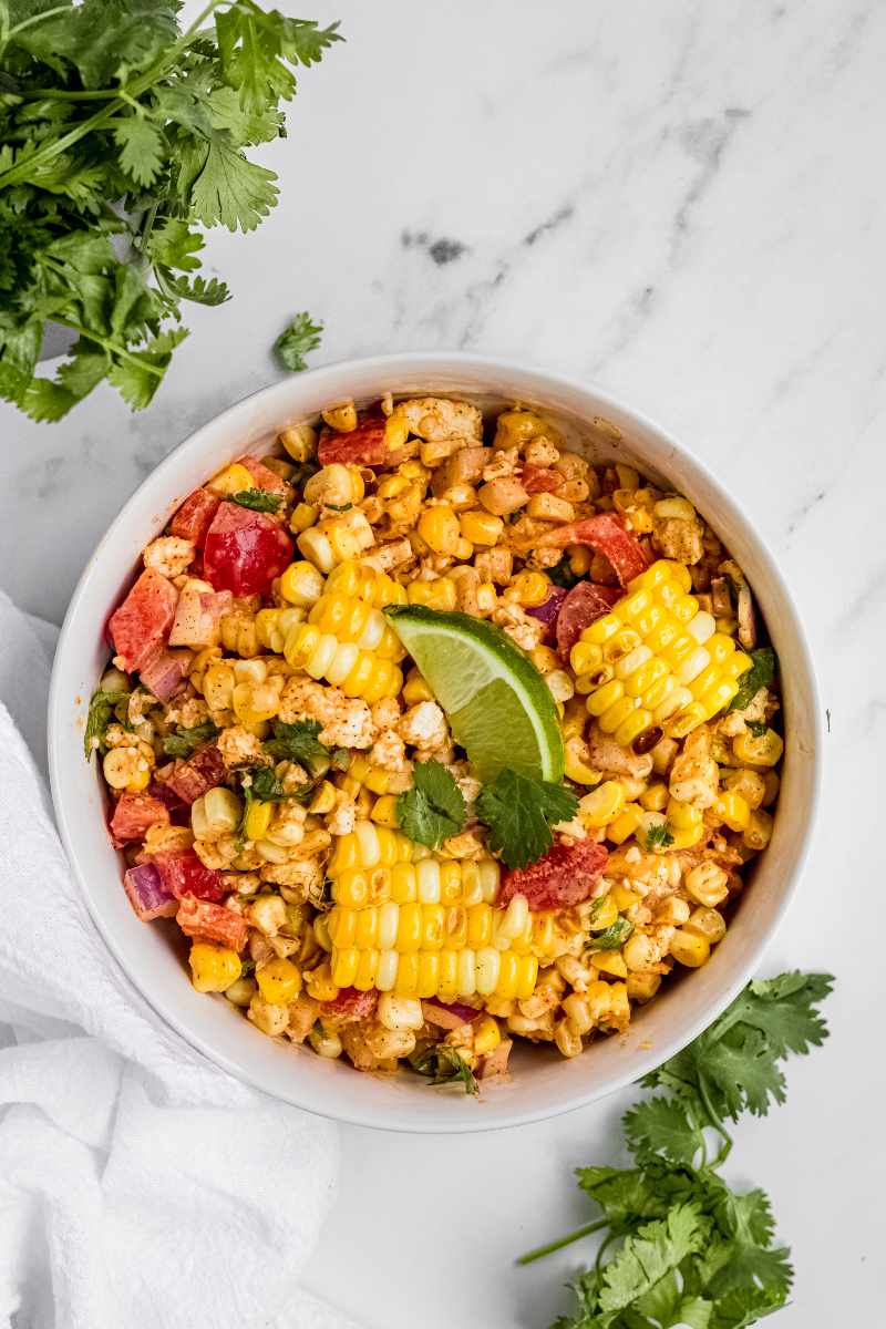 A serving bowl of finished mexican street corn salad garnished with cilantro leaves and a lime wedge.