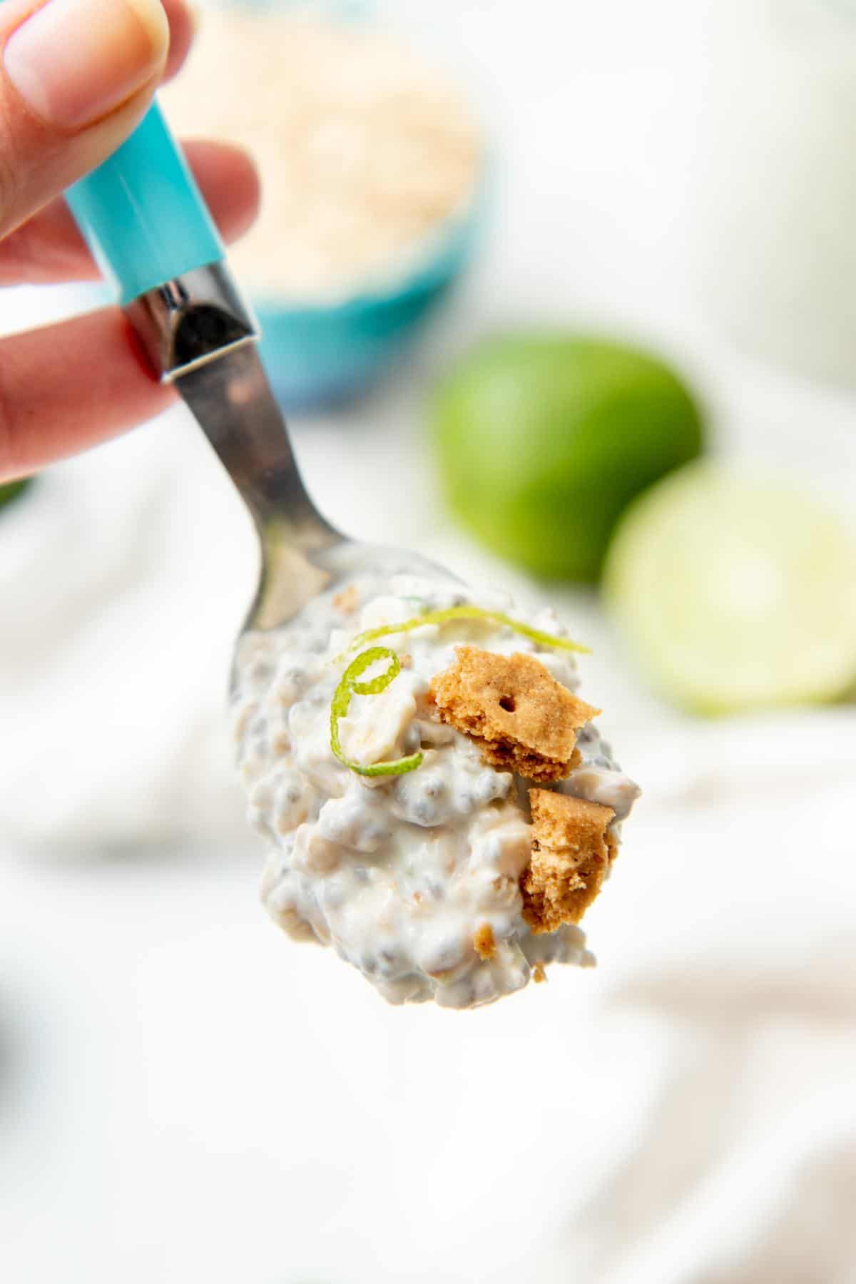 A hand holds a spoon with a blue handle. Spoon is full of overnight oats topped with lime twist and graham cracker crumbles.