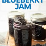 A long spoon scoops up fruit jam from a half-pint jar, fresh fruit and more jars around it. A text overlay reads, "How to Can Blueberry Jam."