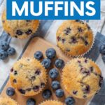 Top view of blueberry muffins on a wooden serving board with fresh berries and a blue and white striped kitchen linen. A text overlay reads, "Blueberry Muffins."