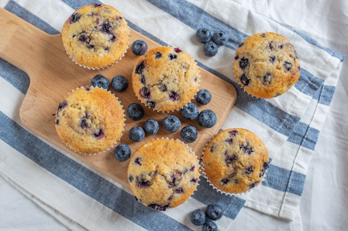 Top view of blueberry muffins on a wooden serving board with fresh berries and a blue and white striped kitchen linen.