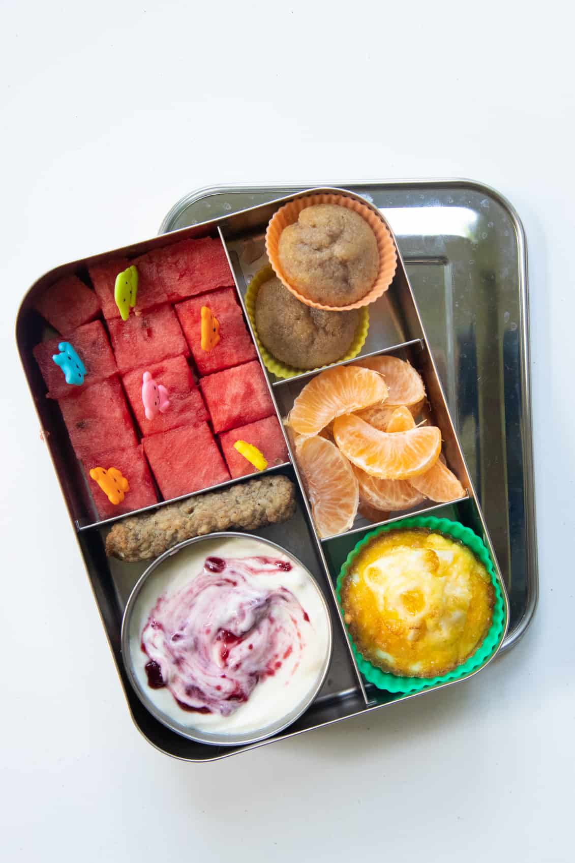 Stainless steel lunchbox packed with brunch foods: watermelon cubes, muffins, egg cup, clementine, yogurt.