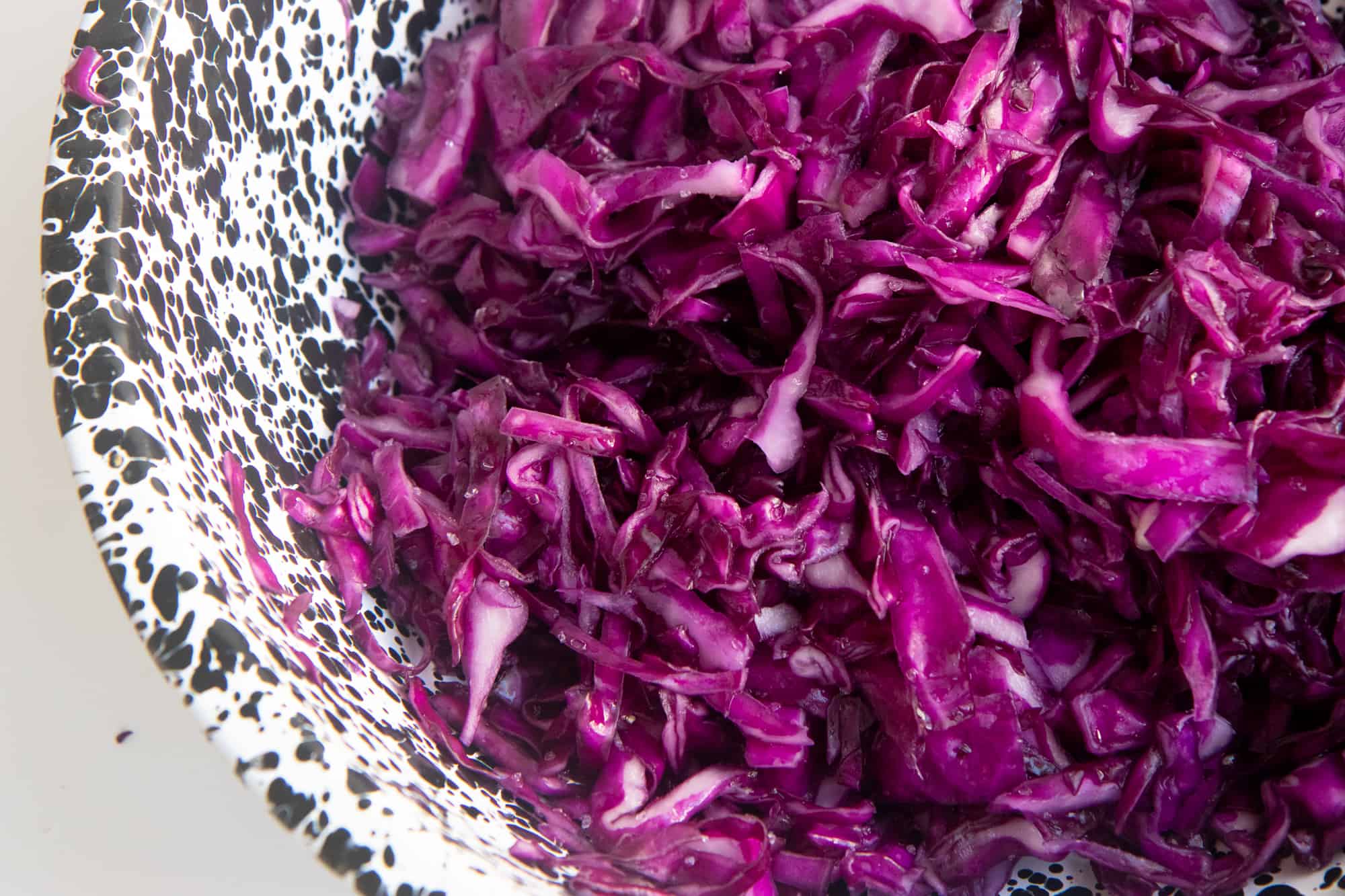 Sliced red cabbage sits in its own brine in a black and white speckled bowl.