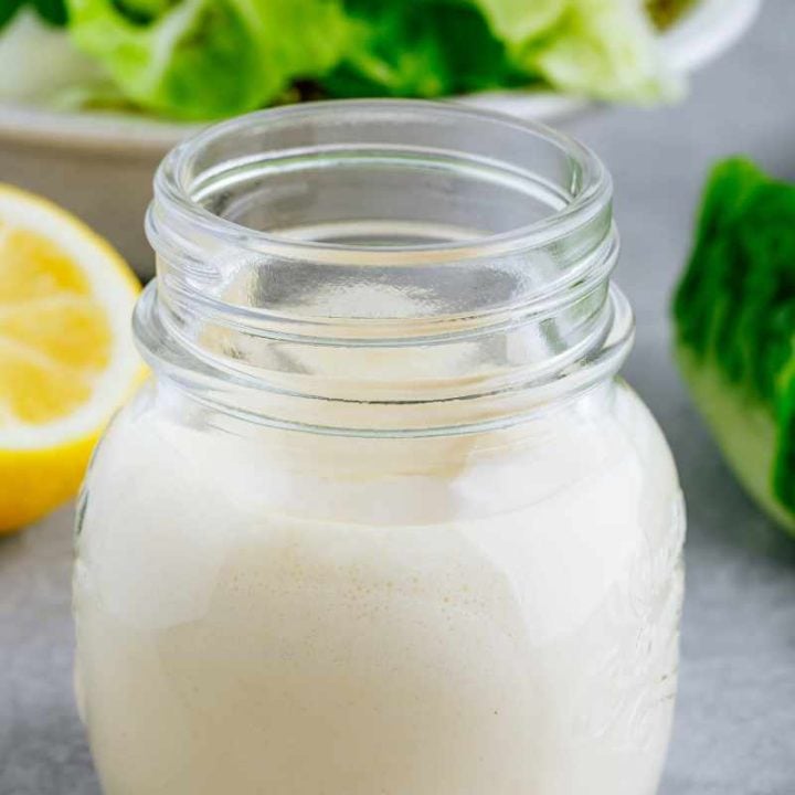 A small glass jar of caesar dressing stands in front of a bowl of crisp romaine lettuce.