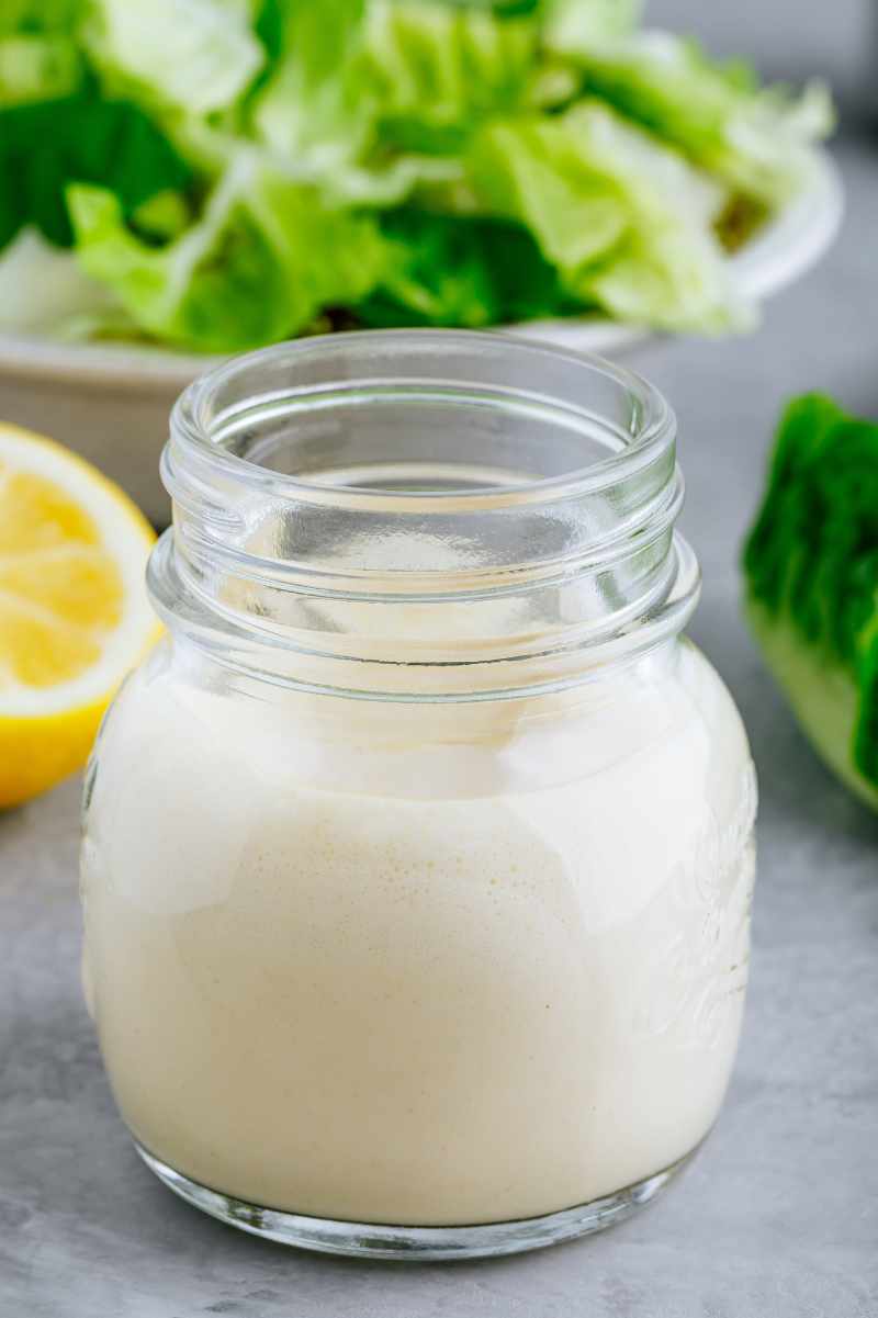 A small glass jar of caesar dressing stands in front of a bowl of crisp romaine lettuce.