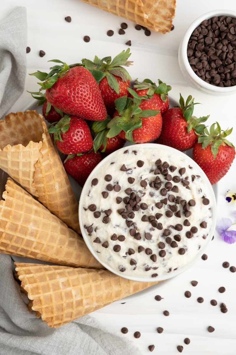 Overhead of a dessert platter with a large white bowl of dessert dip and fresh strawberry and waffle cone dippers around it.