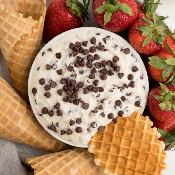 Close view of a waffle cone piece dunked into a bowl of cannoli dip, more waffle cones and fresh strawberries arranged around it.