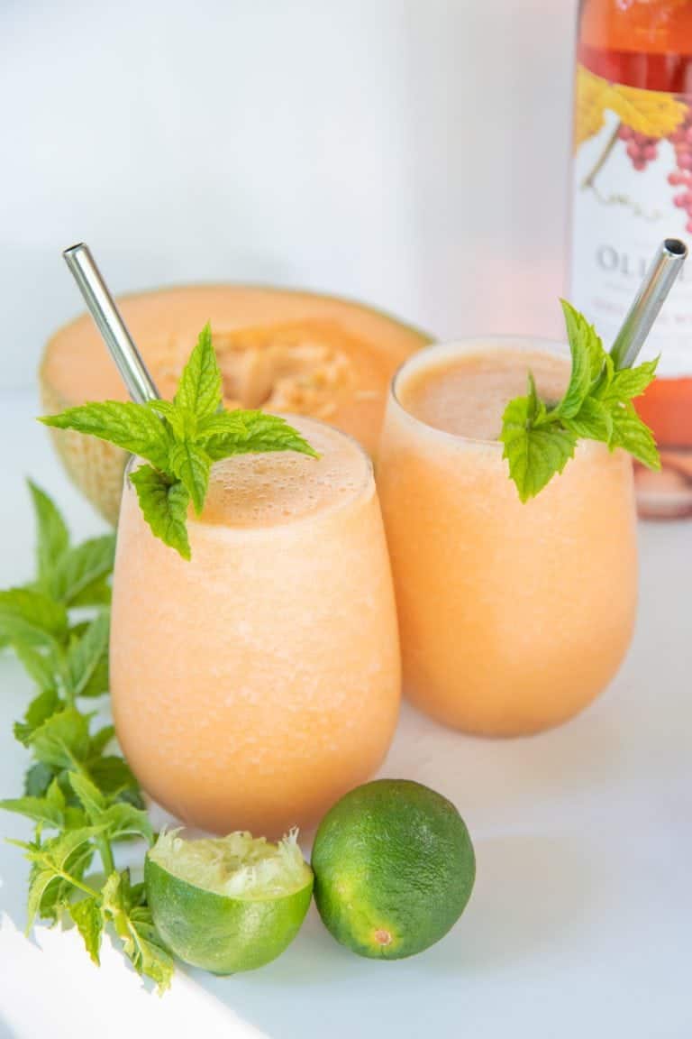 Two glasses filled with cantaloupe frosé, garnished with mint and straws. A halved cantaloupe and mint sprigs surround the glasses.