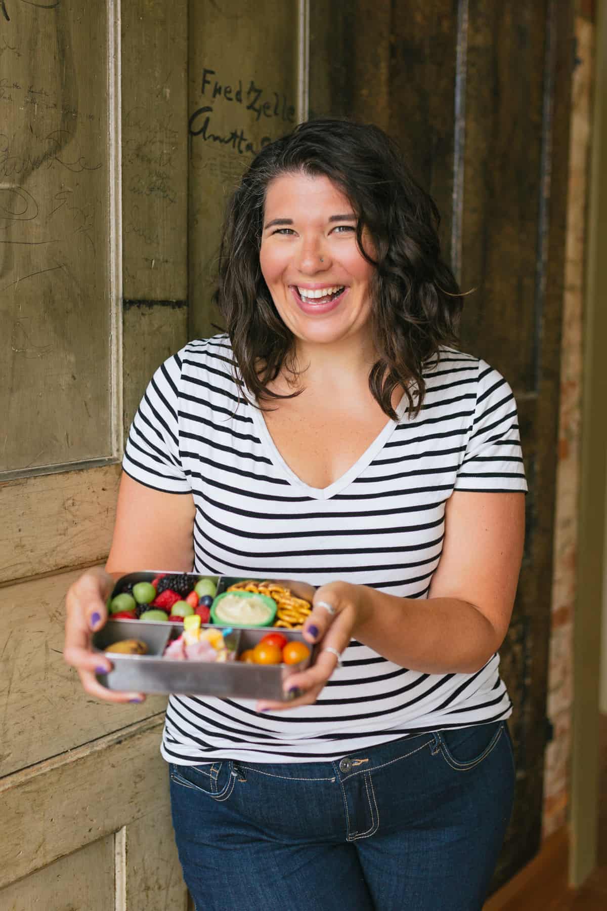 Brunette woman in a striped shirt holding a bento-style lunch box and smiling.