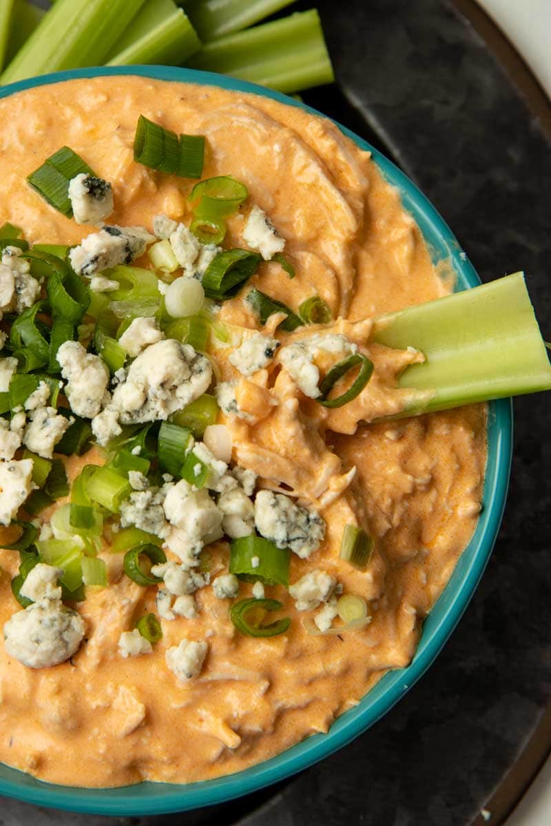 A celery sticks rests in a bowl of spicy, creamy chicken dip garnished with blue cheese and chopped scallions.