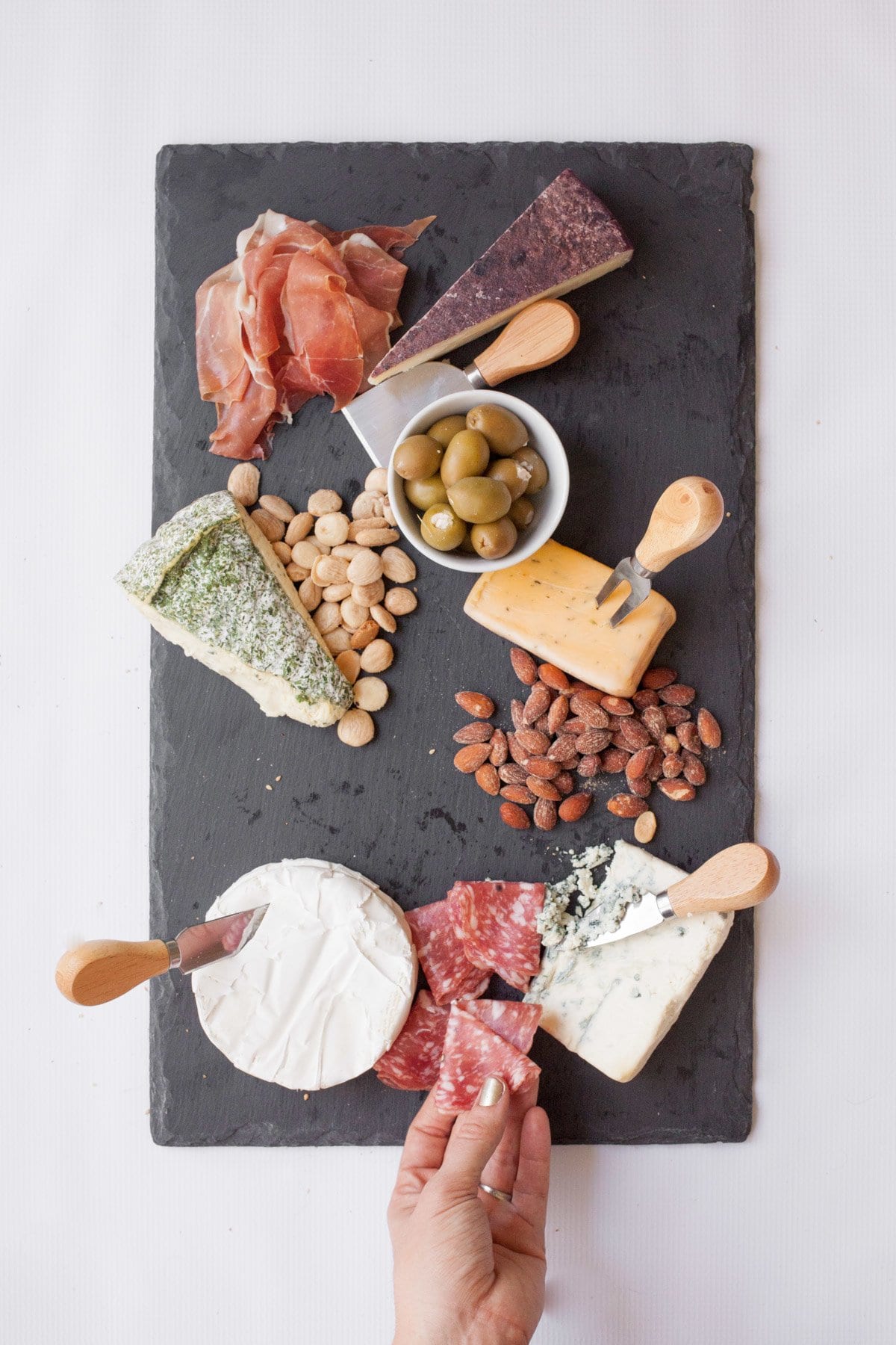 Slate cutting board with 5 wedges of cheese, each with a cheese knife. Various nuts and charcuterie are around the cheese.