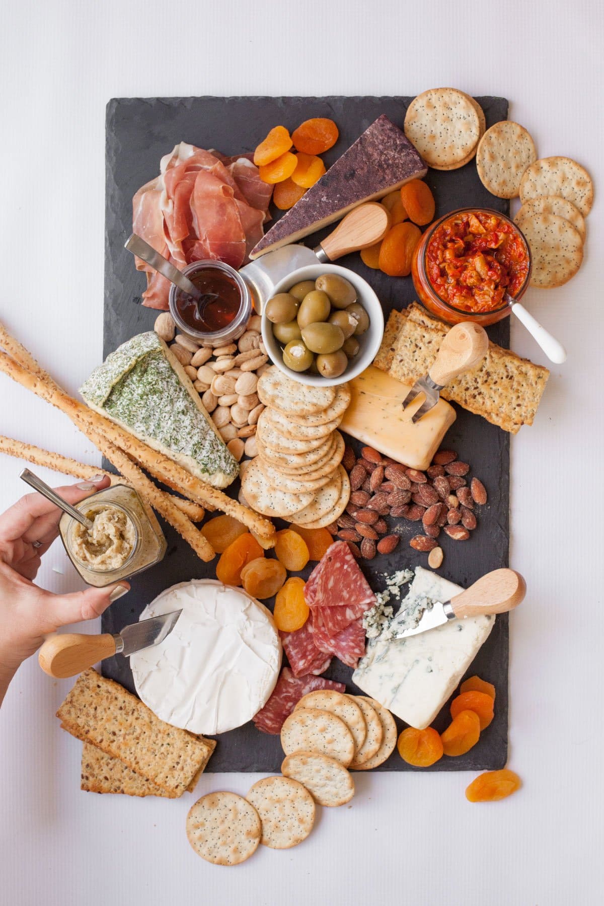 Slate cutting board with 5 wedges of cheese, each with a cheese knife. Various crackers, nuts, dried apricots, olives, condiments, and charcuterie are around the cheese.