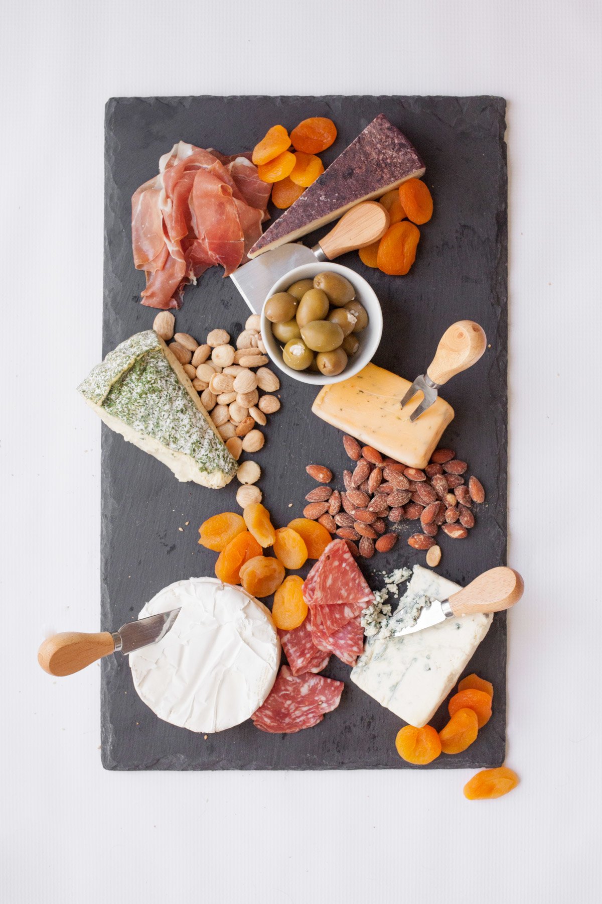 Slate cutting board with 5 wedges of cheese, each with a cheese knife. Various nuts, dried apricots, olives, and charcuterie are around the cheese.