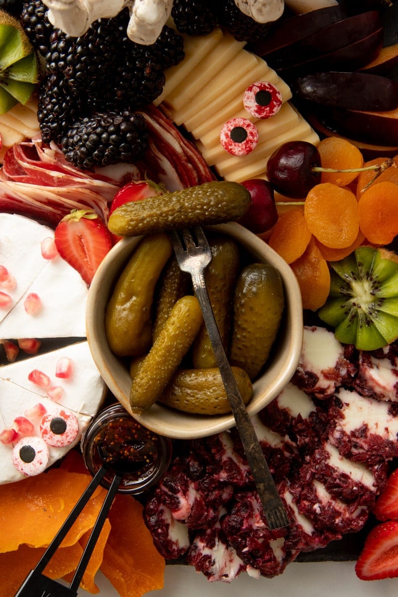 A cocktail fork spears a gherkin atop a bowl of gherkins surrounded by cheeses, meats, fruits, and spooky decor on a cheese board.