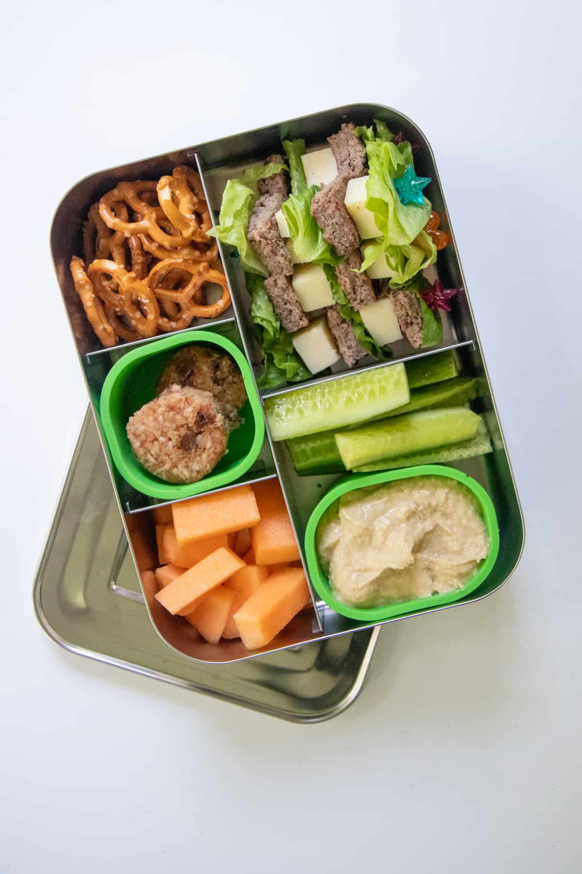 Stainless steel lunchbox filled with a school lunch idea: cheeseburger skewers with melon, pretzels, vegetables and hummus, and cookies