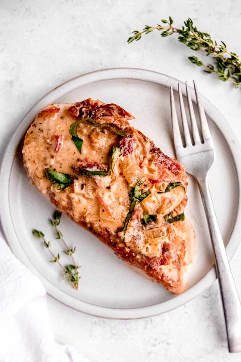 Overhead of a flavorful chicken breast on a light grey plate with a fork and a thyme sprig alongside it.