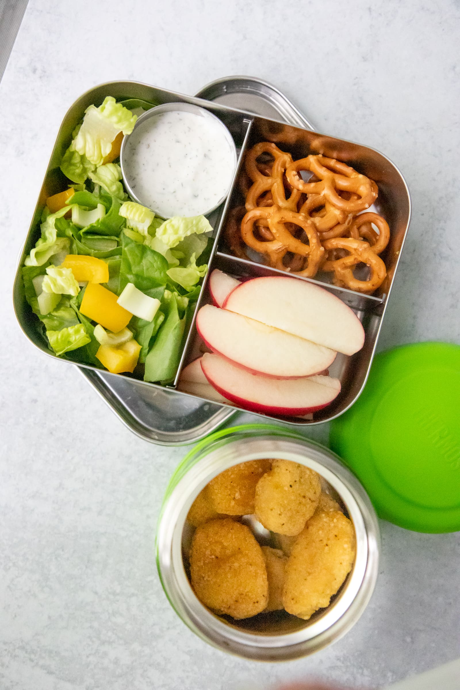 Chicken nuggets in a Thermos, next to a lunch box filled with salad, pretzels, and apple slices