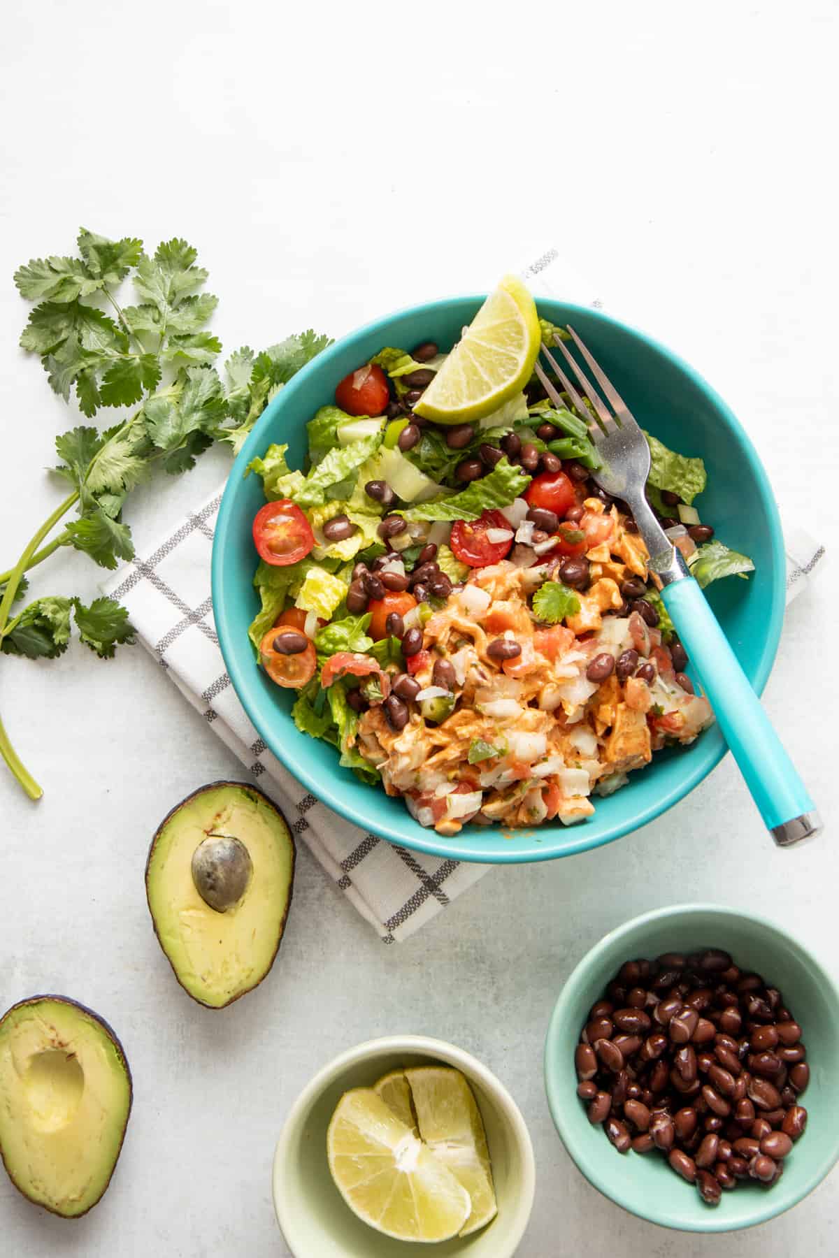 A teal bowl is filled with chicken taco salad. A lime wedge and a fork with an aqua handle rest in the bowl. A halved avpcado rests on a white countertop.