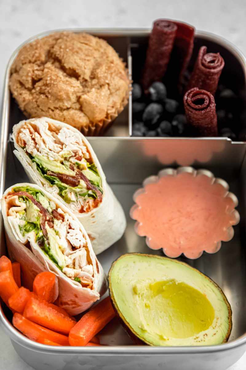 A chicken wrap packed into a metal bento-style lunchbox with a muffin, blueberries, fruit leather, carrots, avocado, and dipping sauce.