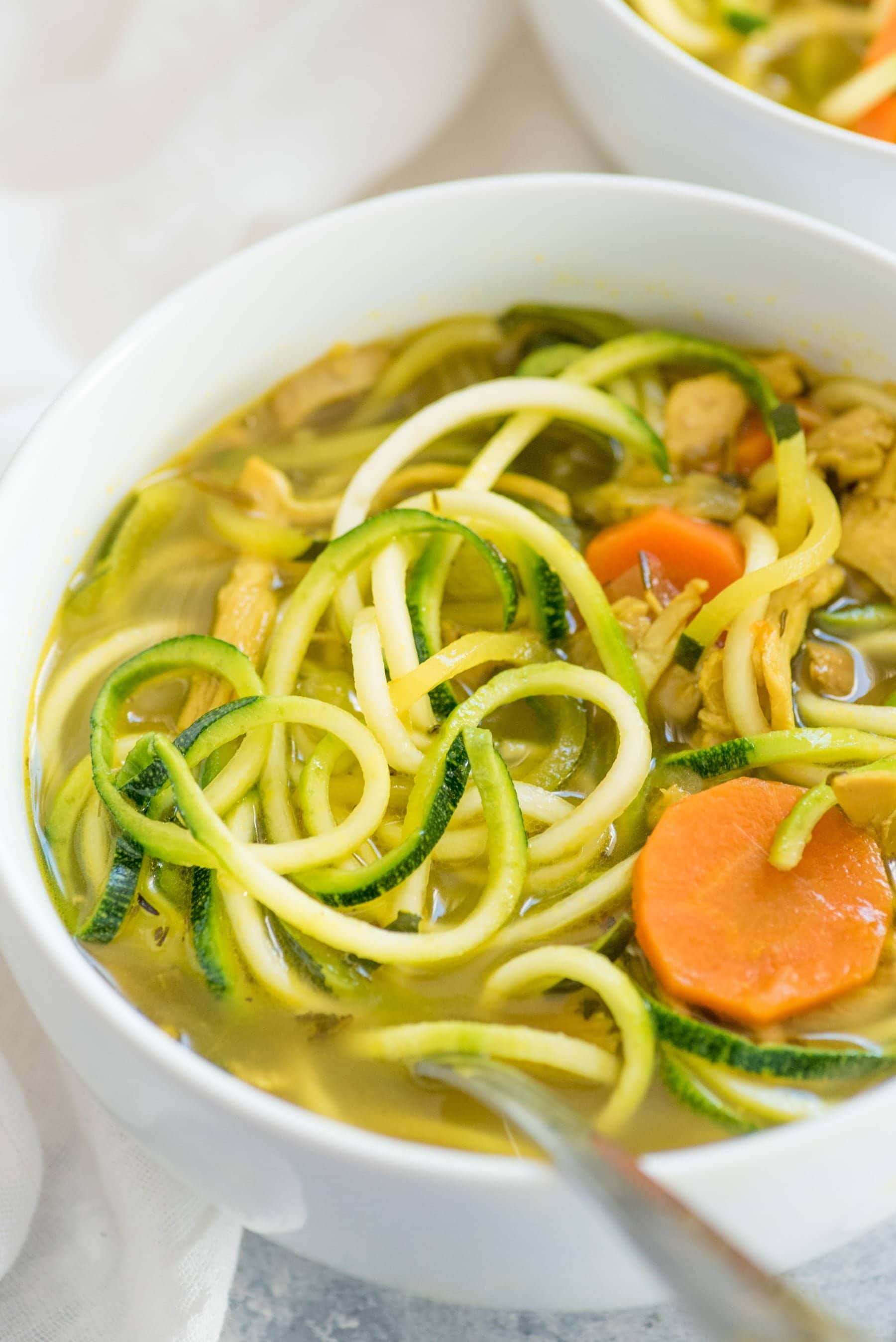 Tight view of Chicken Zoodle Soup in a white bowl with a spoon in the soup. 