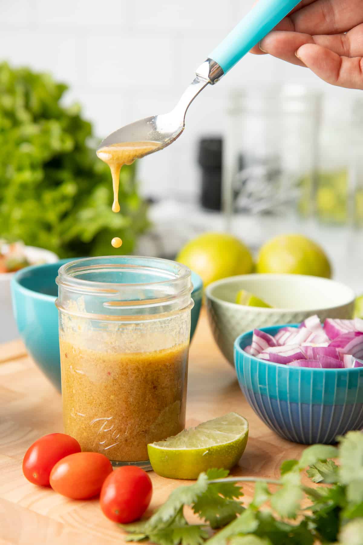 A hand holds a spoon with teal handle. The spoon drips chile-lime salad dressing into a small jar. The jar is surrounded by small tomatoes, a lime slice, and red onions.