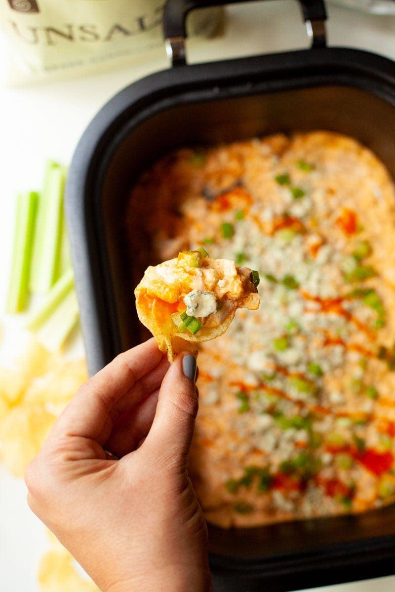 A hand hold up a chip loaded with crackpot buffalo chicken dip. There is celery and chips in the background