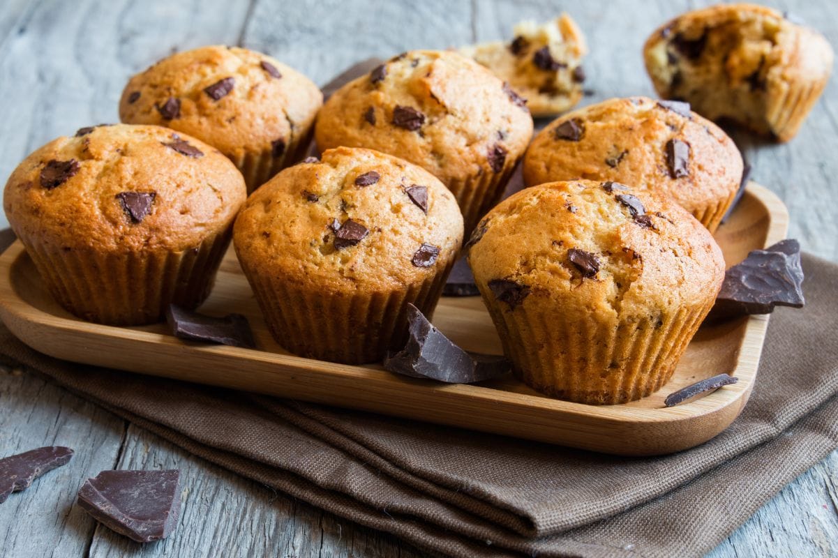 Six chocolate chip muffins on a wooden serving tray with large pieces of broken chocolate around.