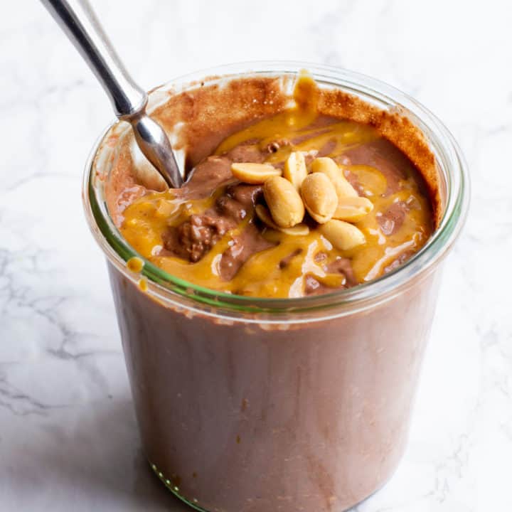 Peanut butter chocolate overnight oats in a glass jar. There are peanuts garnishing the top, and a spoon dips into the jar.