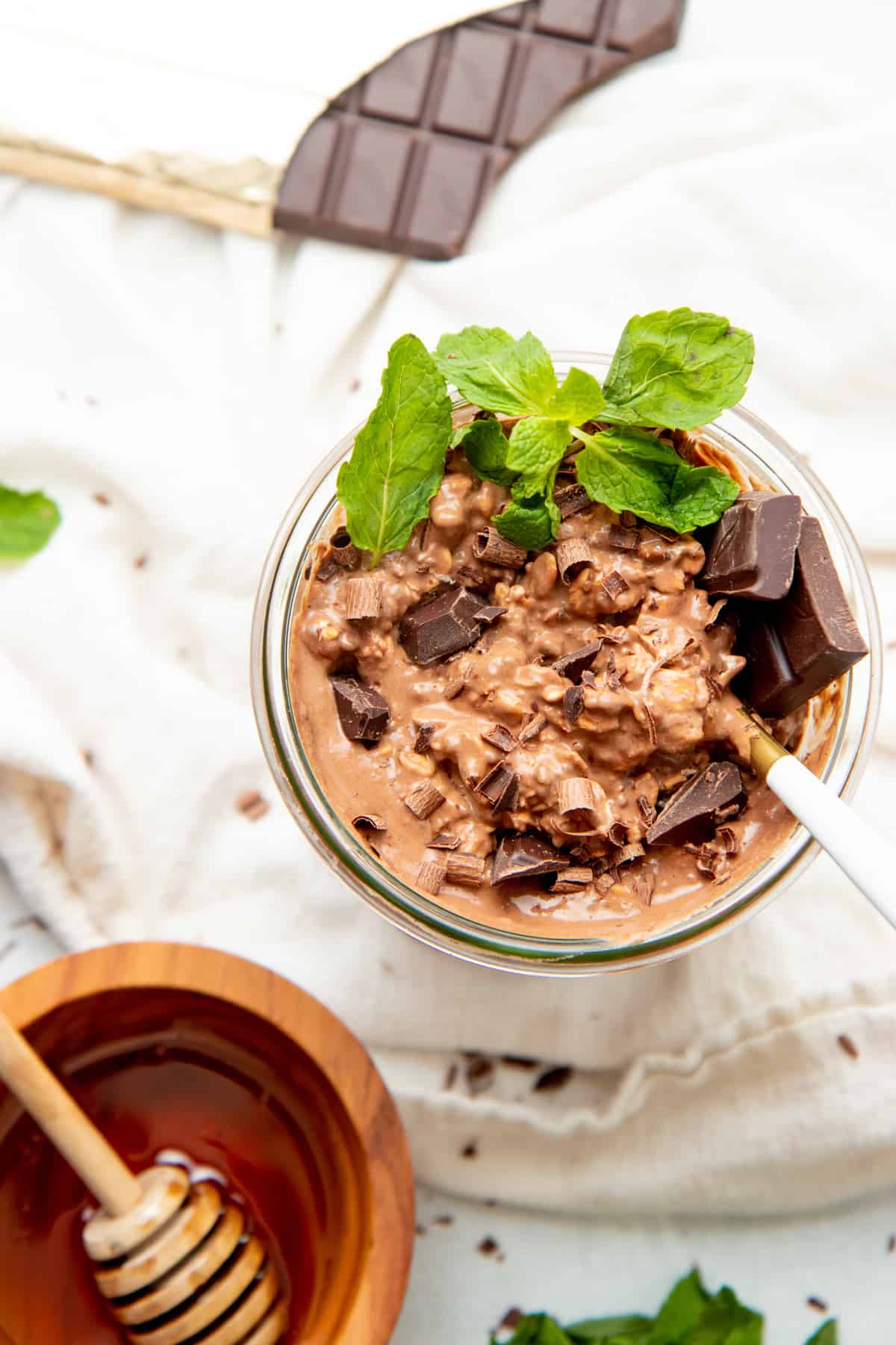 Chocolate peppermint overnight oats fill a glass jar. A small bowl of honey and an opened chocolate bar surround the jar.