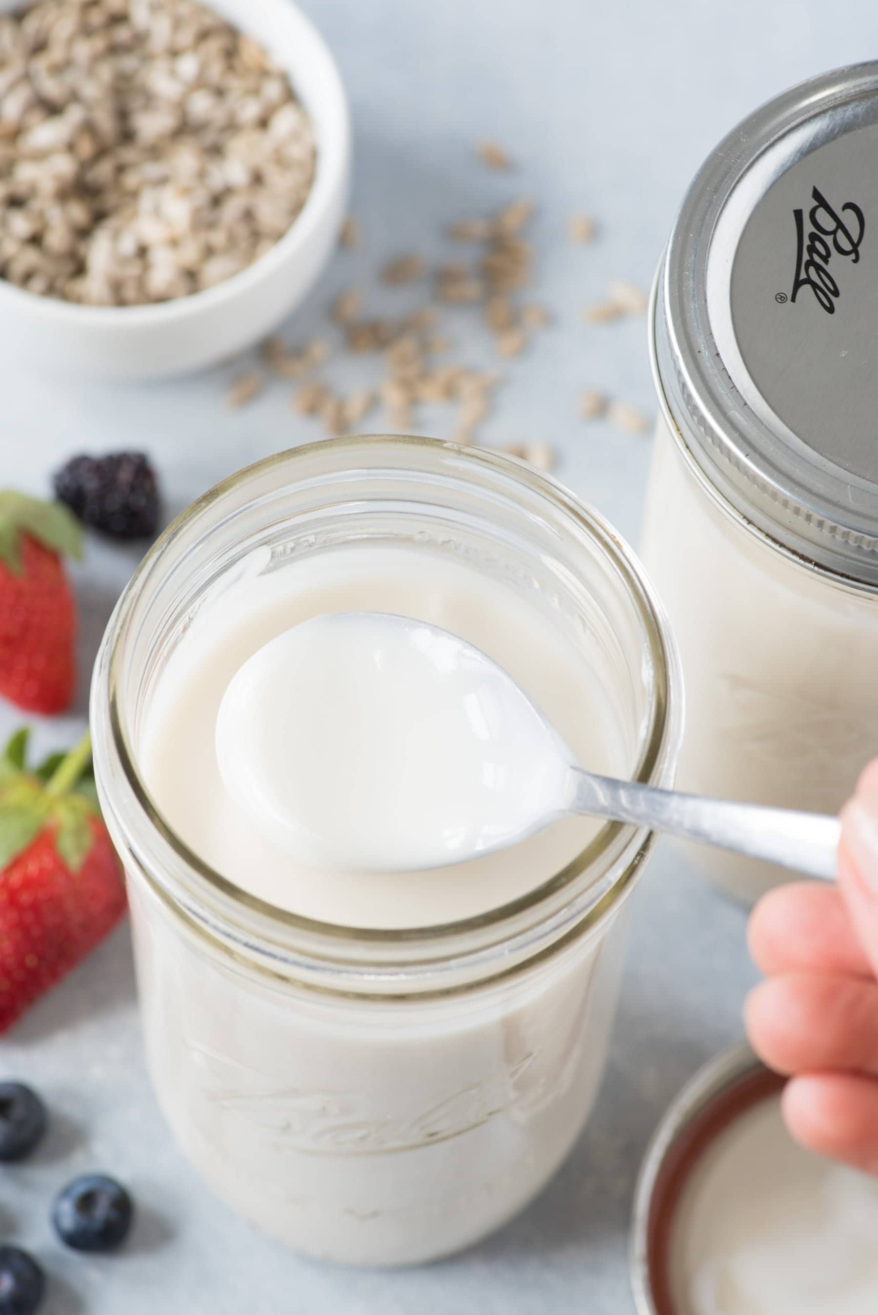 Overhead shot of a spoon scooping Yogurt out of a Ball mason jar, with another jar, berries, and seeds in the background