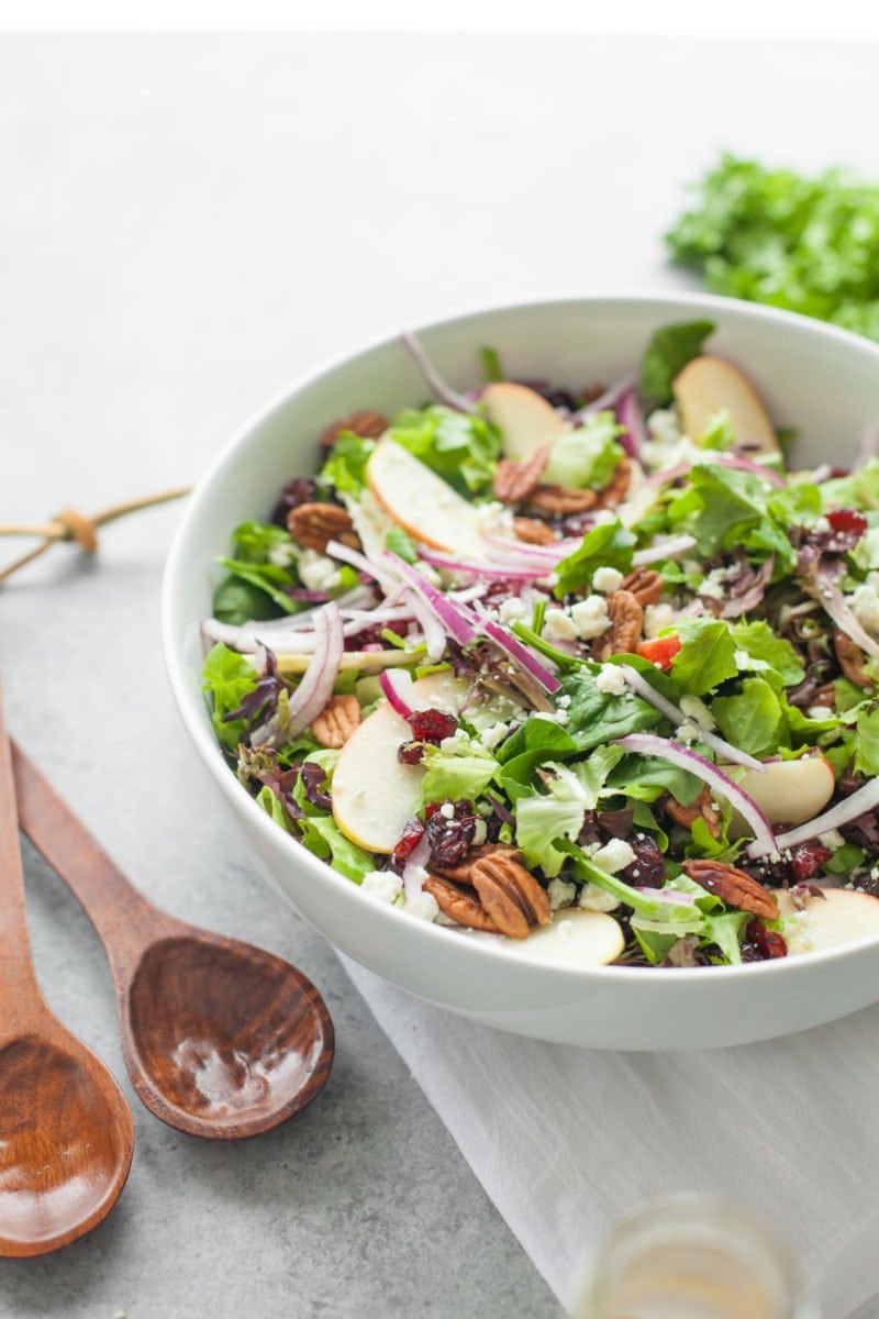 A green salad topped with dried cranberries, toasted pecans, and apple slices sits in a white serving bowl with two wooden serving spoons beside it.