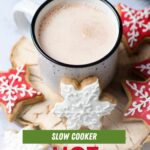 Enamelware mug of crockpot hot chocolate. Red and white decorated cookies surround the mug. A text overlay reads, "Slow Cooker Hot Chocolate."