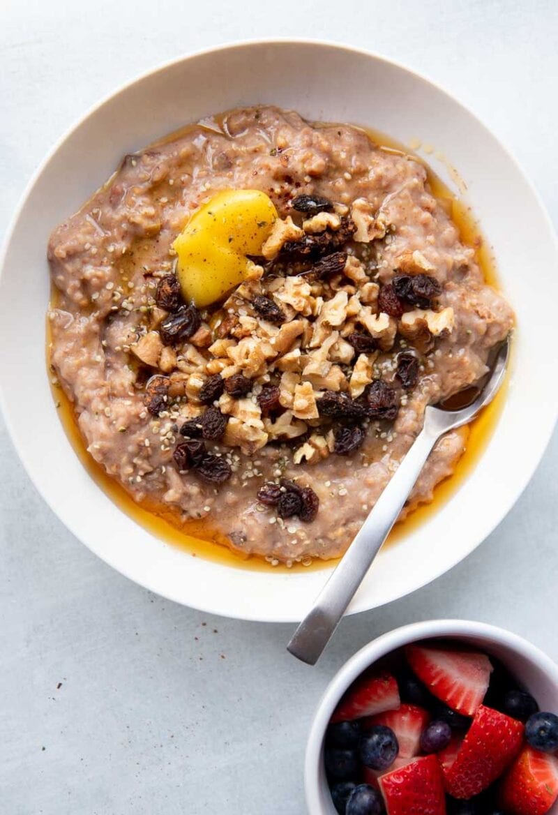 A spoon tucks into a bowl of crockpot oatmeal topped with dried fruit and nuts. A small bowl of berries sits nearby.