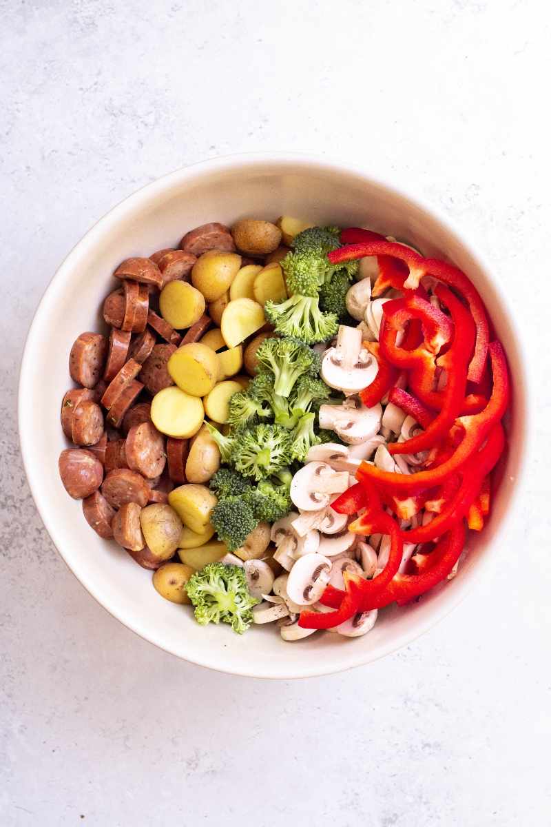 A large white bowl with rows of bite size pieces of chicken sausage, baby potatoes, broccoli florets, mushrooms, and red bell peppers.