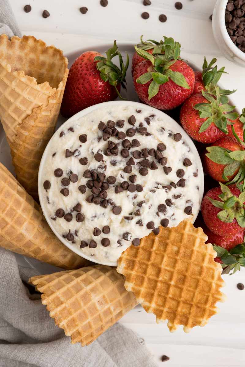 Top view of a platter of dippers, including strawberries and waffle cones, arranged around a bowl of creamy dessert dip topped with mini chocolate morsels.