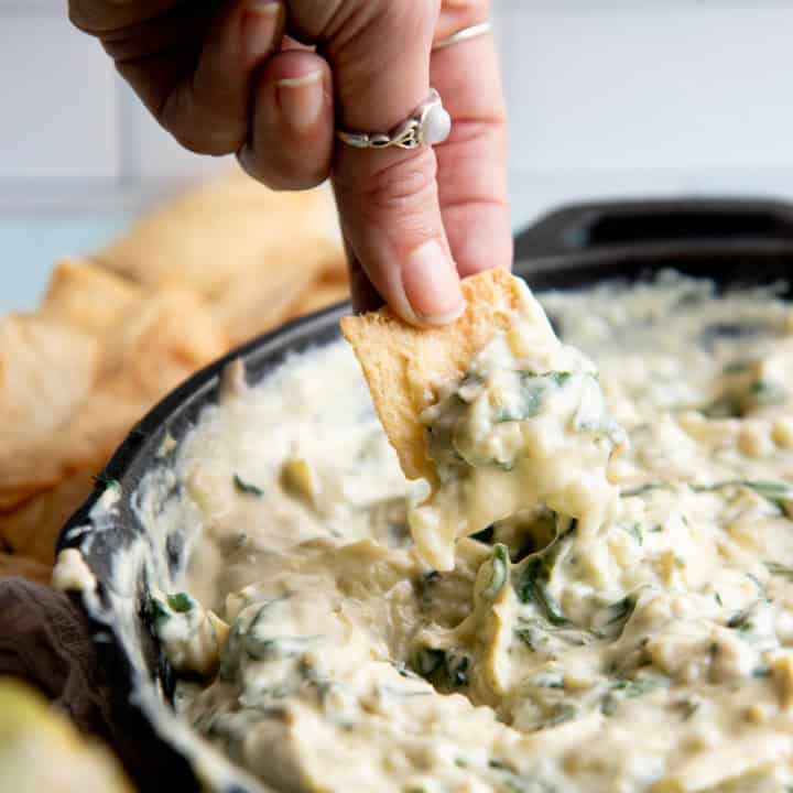 A hand dips a pita chip into a skillet of vegan spinach artichoke dip.