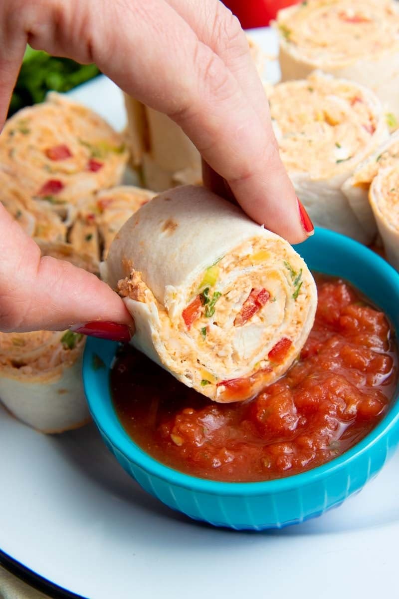 A hand dips a taco pinwheel into a small bowl of salsa.