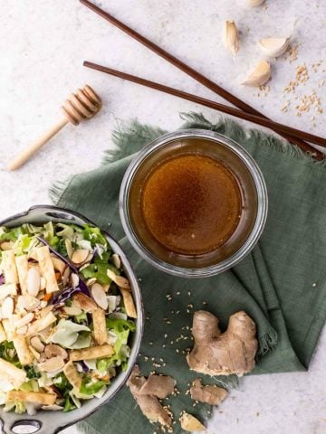 Overhead of sesame ginger dressing in a bowl beside a bowl of salad, fresh ginger, garlic, and sesame seeds around them.