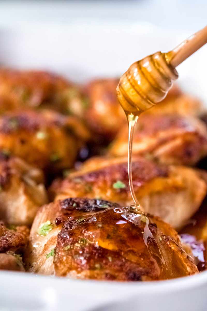 A honey dipper drizzles honey onto a baked sticky chicken thigh.