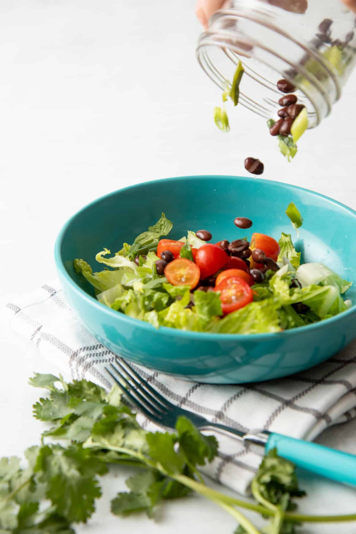 A hand pours a salad from a jar into a teal bowl. The bowl sits on a white and brown plaid dishtowel. A fork with a teal handle sits to the side of the bowl.