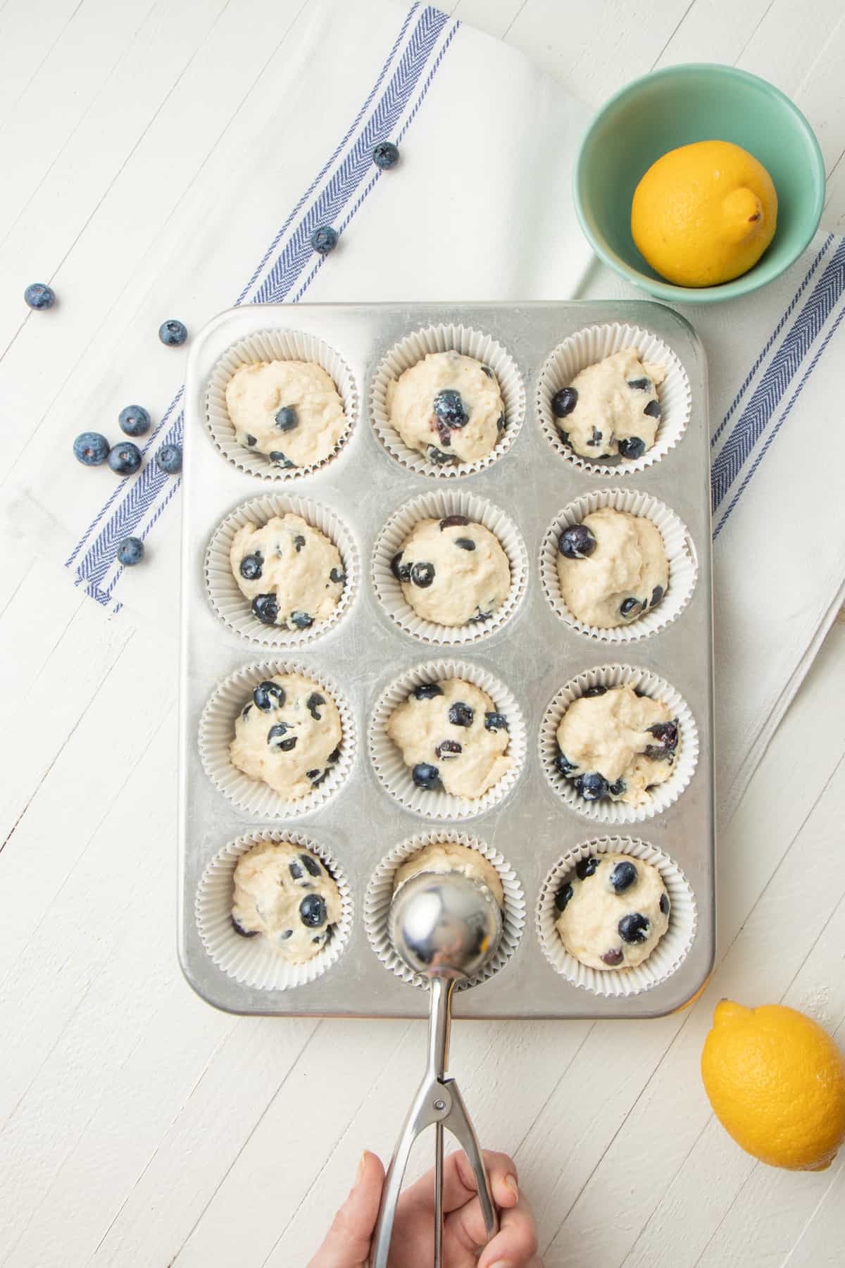 A hand uses a dough scoop to fill a muffin tin with batter.